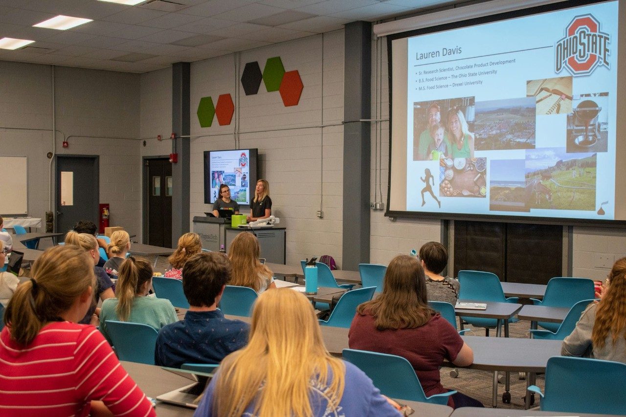 Two presenters speak to a full classroom.