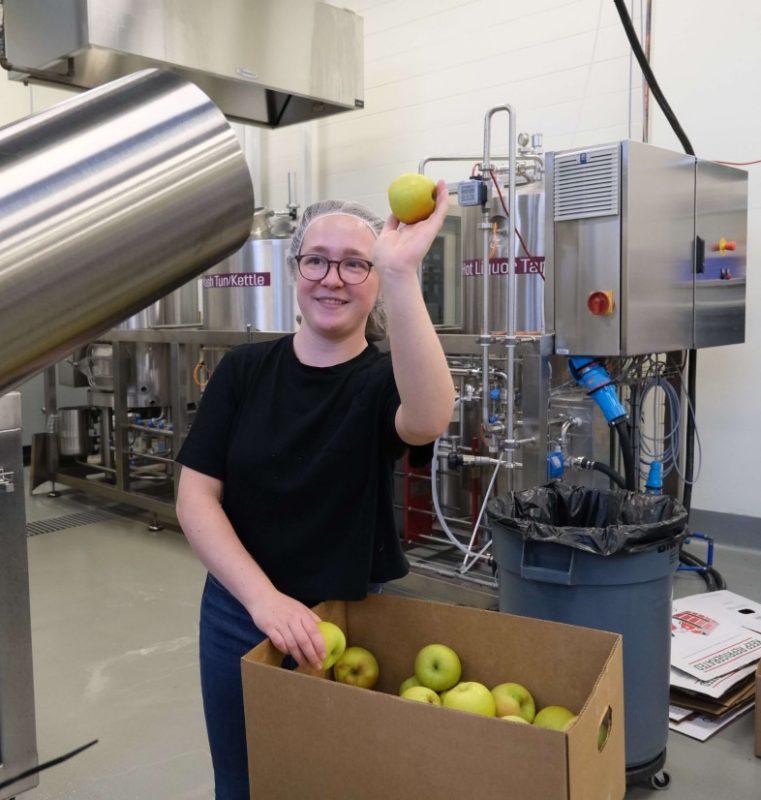 A person holds up an apple while standing next to a box of apples.